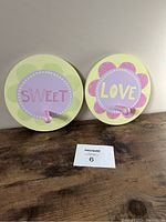 Two round decorative wall hooks side by side on a wooden surface showing the front with pastel floral designs and words 'SWEET' and 'LOVE'.