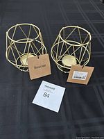 Front view of two gold wire geometric candle holders placed on a black surface, showing Bouclair tags attached.