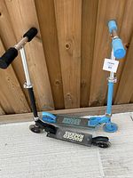 Two kids scooters, one black and one light blue, shown side by side against wooden background.
