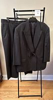 Full view of the dark grey men's suit jacket and pants displayed on hangers and a rack.
