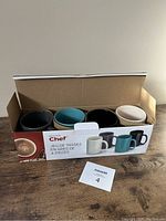 Open box showing four stoneware mugs of different colors: black, dark teal, dark brown, and off-white.