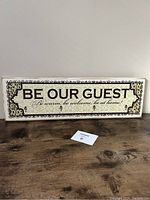 Front of rectangular wooden sign with 'Be Our Guest' message and decorative corners