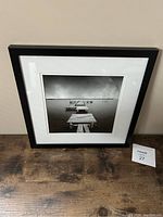 Front view of the framed black and white photo depicting a wooden dock over calm water, resting on wooden surface against a beige wall.