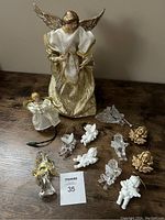Photo showing a large angel tree topper with white and gold robes, a smaller white angel tree topper with gold trim, clear glass angel ornament, multiple white angel figurines in different styles, clear plastic angel ornaments, and gold-tone angel rose-shaped ornaments arranged on a wooden surface.