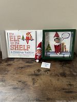 Front view of boxed Elf On The Shelf Boy Edition figure with blue eyes, extra elf figure, and large book display