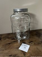 Front view of the glass drink dispenser showing embossed branding, metal lid, and spigot on wooden surface.