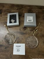 Two Mikasa crystal round ornaments shown with original silver boxes and hanging ribbons on wooden surface. One box shows product barcode and labeling.