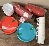 Photo showing 6 white plastic bowls stacked on left, two sets of 4 plastic plates (one red, one blue) in middle, 6 white plastic textured cups stacked on right side, and two rolled red blankets in carrying bags behind the plates and bowls.