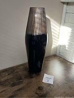 Photo of tall brown vase with etched vertical lines near top, placed on wooden surface with sunlight from side.