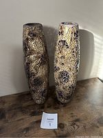 Photo showing two tall mosaic vases placed side by side on a wooden surface against a wall, highlighting their colors and mosaic design.