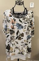 Front and side view of white blouse with floral print displayed on a black metal mannequin, showing short sleeves and loose fit.