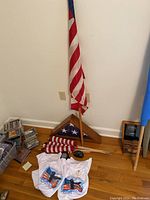 Photo showing folded large American flag in wooden display case, several folded small American flags with wooden poles, folded patriotic T-shirts with eagle and flag design, and handheld tape measure on wooden floor.