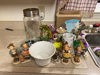 Five Hummel figurines, white bowl, Ball mason jar, ceramic tea dispensers, decorative items and marbles on kitchen counter