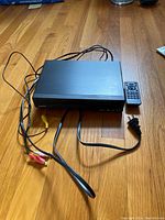 Black Kenwood CD player with attached cords and remote control on wooden floor.