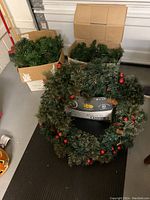 Artificial decorated Christmas wreath with red baubles and pine cones in foreground; three boxes of green artificial garlands behind it in garage.