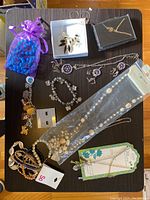 Photo shows various items on a table: purple pouch with blue beads, silver leaf brooch/pendant in a box, gold bird pendant necklace in a box, silver-tone chain necklace with purple charms, crystal bracelet, white bead necklace in packaging, assorted beaded bracelets with shell charms.