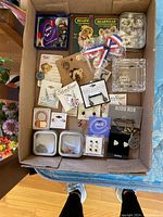 Top-down view of a box containing assorted costume jewelry including earrings, pins, and jewelry cases. Various brands and styles noted.