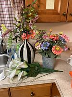 Photo showing three faux flower arrangements on kitchen counter: black and white vase with faux flowers, galvanized bucket with colorful flowers, and white calla lilies bundle.
