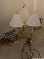 Photo showing three brass lamps with white pleated shades. Two lamps are shorter with identical styling and one is taller with a finial on top. All have cords and inline switches.