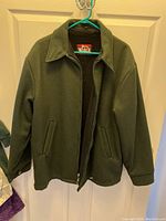 Front view of men's Woolrich coat hanging on door, showing green wool material, collar, zipper, and side pockets