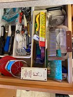 Top view of first kitchen drawer showing onion holder, garlic press, plastic slicers, scissors, and various small utensils in compartments.