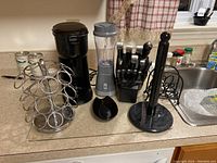 Wide shot showing all items arranged on a kitchen counter: black coffee maker, blender, knife set, paper towel holder, metal cup holder.