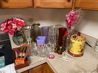 Various glass and ceramic vases, wooden and glass candle holders, a ceramic candle warmer, artificial pink flowers, and decorative marbles arranged on a kitchen countertop.