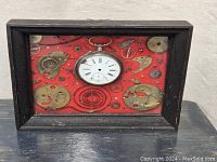 Front view of framed artwork showing black shadow box frame enclosing a pocket watch face and assorted watch parts on red background.