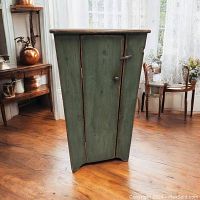 Front angle view of the rustic pine cupboard showing tapered shape, green wash finish, single door with knob and latch.