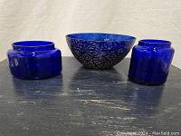 Front view of two lidless cobalt blue glass jars and a cobalt blue glass bowl with black crackle design on a dark table against beige background.