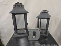 Two black cast iron lanterns and a galvanized metal letter D arranged on a table against a neutral backdrop.
