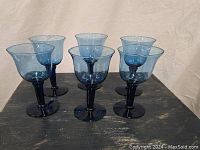 Set of six vintage cobalt blue hand blown wine glasses arranged on a dark wooden surface against a fabric backdrop. Glasses are shown upright, highlighting their blue stems and bowls.