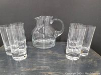 Photo showing the water pitcher centered with six tall glasses arranged around it on a dark wooden surface, some glasses are labeled.