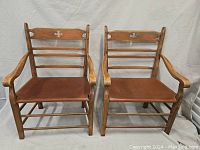 Two wooden arm chairs side by side with tan leather sling seats and horizontal slats with decorative cutouts on backrests.