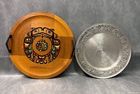 Photo showing two trays side by side: left wooden tray with colorful Native First Nations art and black handles, right metal tray with embossed border design
