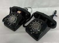 Two black rotary dial telephones side by side on white background, visible rotary dials and handsets