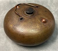Top perspective showing round form, engraved floral pattern on bronze surface and hinged bail handle resting across top