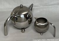 Photo showing the stainless steel teapot with lid and matching creamer on white background, highlighting their atomic spherical design and reflective finish.