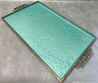 Top side angle view of rectangular metal tray showing textured aqua moiré glaze surface, brass frame, and decorative cut-out metal handles on a grey surface.