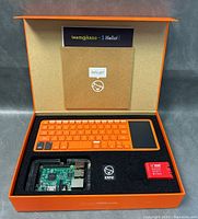 Open box showing orange keyboard with integrated touchpad, Raspberry Pi board in case, Kano USB drive and red Wi-Fi dongle securely fitted in black foam insert.