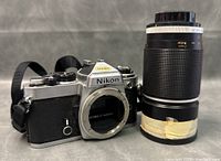 Front and body of Nikon FE camera showing lens mount open, attached strap and Panagor lens standing upright