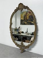 Overall view of the full oval Louis XVI style mirror showing its ornate gold-tone metal frame with shell motifs at top and bottom, leaning against a white wall.