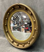 Framed oval convex mirror angled to show front and side details, gold frame with decorative round protrusions.