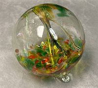 Clear glass orb with colorful green, orange, yellow, red internal swirls, resting on three small glass feet. Minor wear visible on surface.