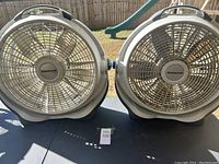 Two large beige box fans branded WindMachine by Lasko, side by side on a table in outdoor setting.