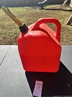 Front view of red 5 gallon plastic gas can with yellow spout and carrying handle, placed outdoors on a table.