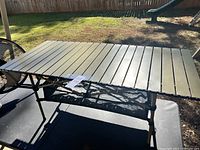 Image of rectangular aluminum camp table set up outdoors on black surface. Shows light olive green slatted tabletop and collapsible brown metal legs with black mesh storage shelf below.