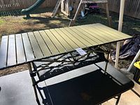 Top-down angled view of an aluminum camping table with slatted tabletop and mesh shelf underneath on a black surface outdoors.