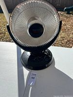 Front view of black heater fan on white surface outdoors, showing circular grille and base with control dials.