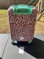 Front view of pink hard shell luggage with black leopard print, green handle and wheels visible on outdoor table.